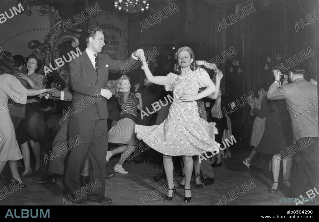 Orig. bildtext... SPRING IN THE STRAND. Screen star ANNA NEAGLE - once a dance hostess - has coached co-star MICHAEL WILDING for a dance sequence in husband Herbert Wilcox's Spring in Park Lane at Elstree. To-day (Wednesday), they joined the dancers at the Lyceum in a boogie-woogie number for filming. PICTURE SHOWS: ANNA NEAGLE and MICHAEL WILDING cut a rug at the Lyceum to-day (Wednesday). Anm. Neagle, Anna, 1904-1986, brittisk skådespelare, professionell dansare vid 14 års ålder, kvinnlig medlem av brittiska Empireorden 1969 Wilding, Michael, 1912-1979, brittisk skådespelare Filmen Spring in Park Lane hade premiär 1948 Klänningar Pardans Blommigt Balans Samspel Vidd Svänga CD404 persons: MICHAEL WILDING;ANNA NEAGLE sites: STORBRITANNIEN;LONDON.