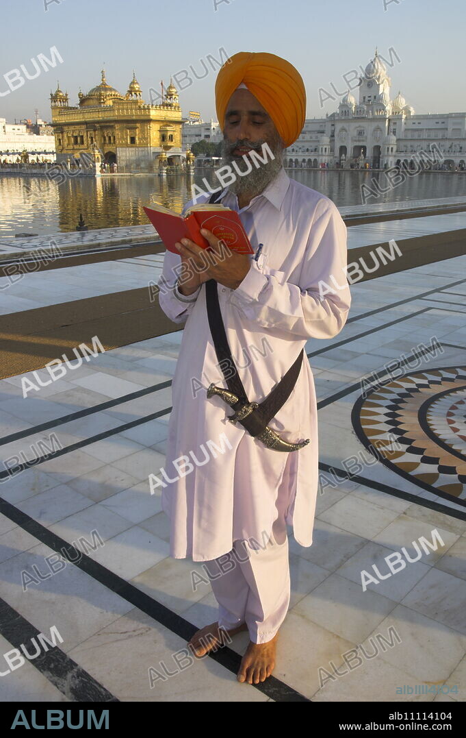 Sikh pilgrim with orange turban, white dress and dragger, reading prayer book, in front of the Golden Temple, Amritsar, Punjab state, India, Asia.