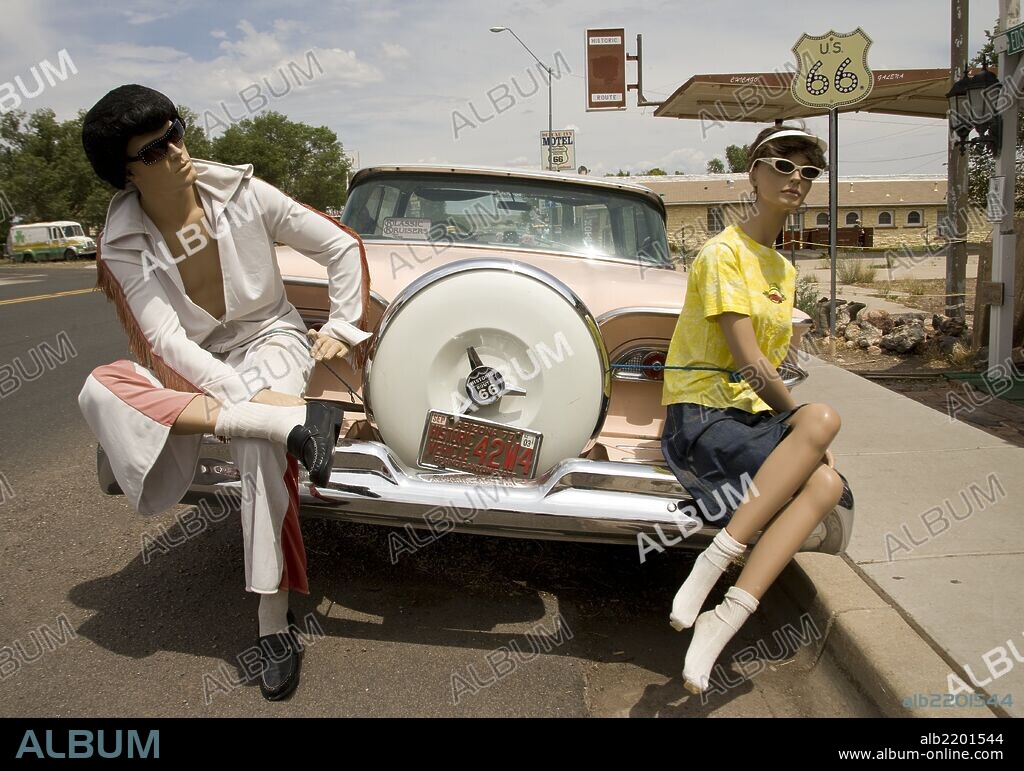 An Elvis Look-alike dummy sits on the rear end of a pink ford aside a tire holder marked Route 66.