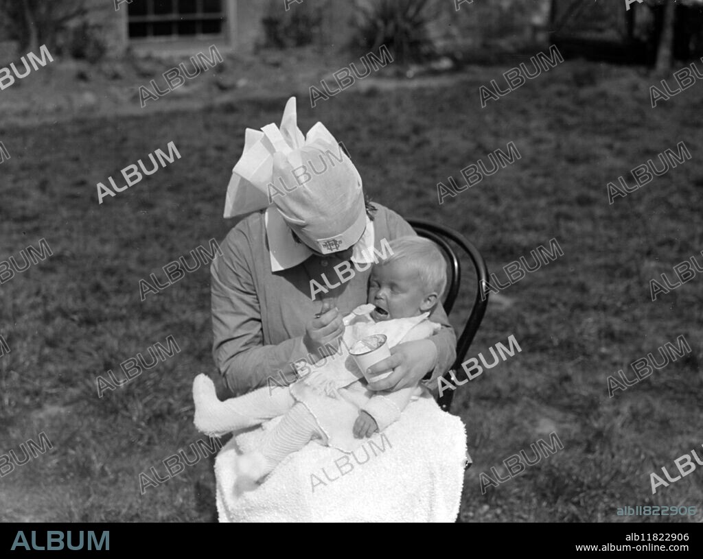 Buxted Lodge, Clapham, London. Children's Nursery babies. A blonde baby sitting on a nanny's knee and being fed. Sitting on a chair in the garden. The baby is wearing wollen tights. possibly 1920s.