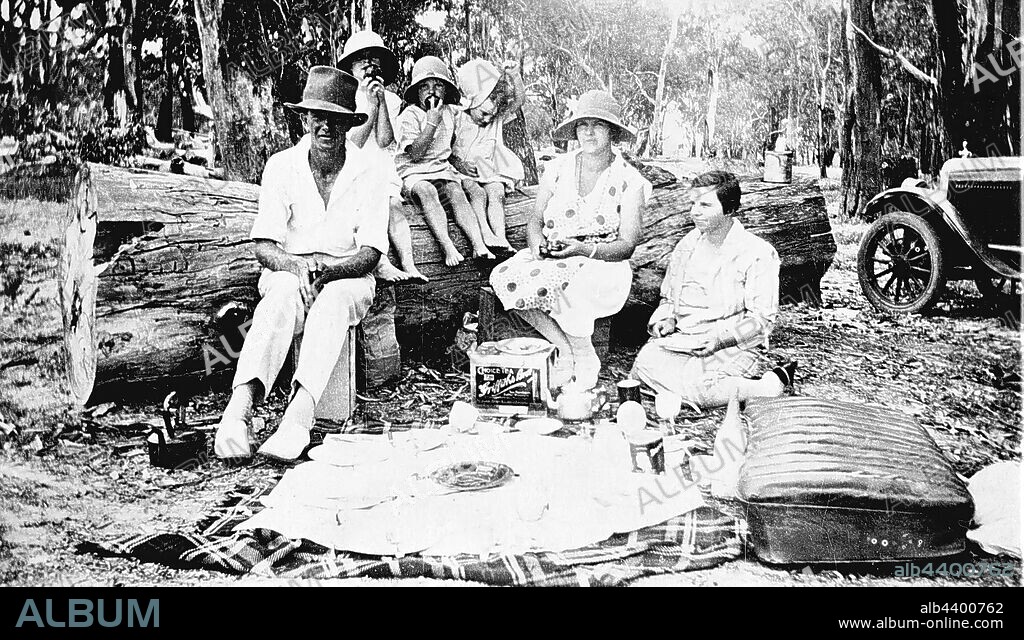 Negative - Victoria, circa 1930, A family at a picnic. The food is on a cloth spread on a rug. The children are seated on a tree trunk and a car seat is on the right.