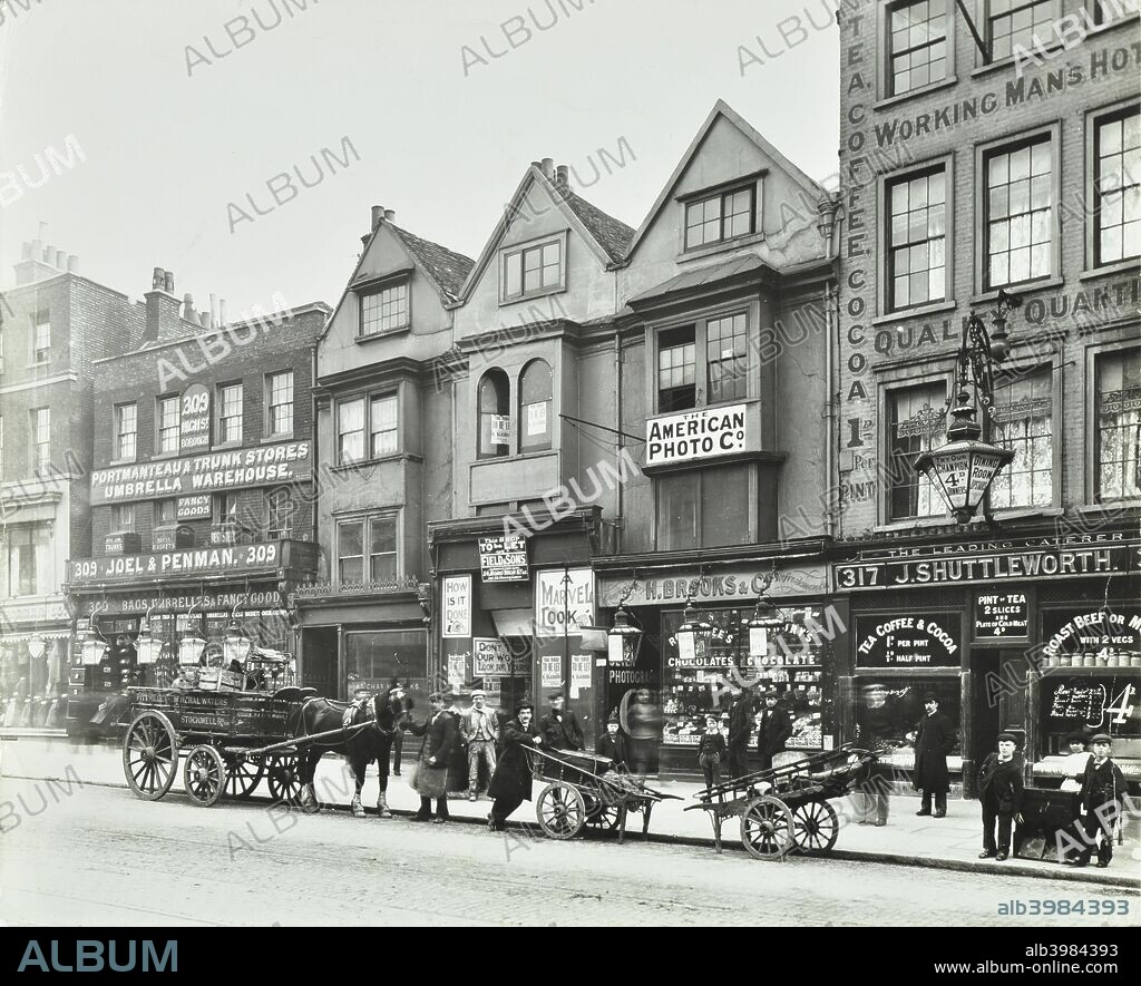 Horse drawn vehicles, barrows and shop fronts, 309-317 Borough High Street, London, 1904. Men and boys wait with barrows and horse-drawn carts. Behind are W Brooks & Co Chocolatiers, Joel & Penman Warehouse and J Shuttleworths Coffee Shop, and a working man's hostel where hot drinks are sold for a penny.