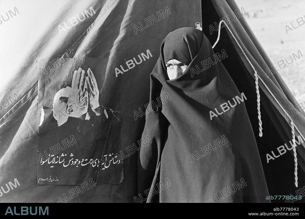 Guerre Iran-Irak (22.09.1980 - 20.08.1988) / Front iranien / Réfugiés. Dans un camp de réfugiés, près de la frontière avec l'Iraq, une très jeune femme portant un tchador se tient près d'une tente sur laquelle est fixée une affiche de l'ayatollah Khomeini en prière, avec des paroles écrites. Pendant les combats, toute la population de la ville de Sarpol-e Zahab dans la province de Kermanshah à l'ouest de l'Iran, à 20 km de la frontière avec l'Irak et à 180 km de Bagdad à été éloignée du front. Photo 12.10.1980.