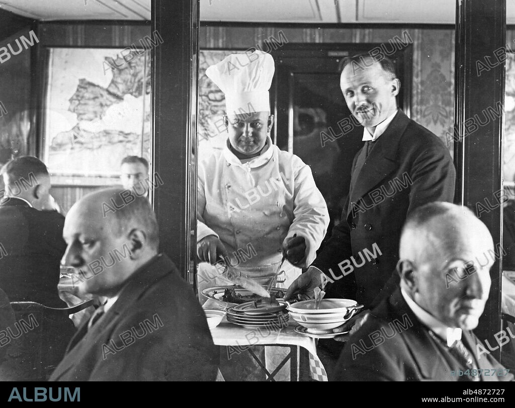 GERMANY 1929. Orig. caption ... FIRST AND ONLY PICTURE REPORT of the great Oriental voyage of the "Graf Zeppelin", taken by our special image reporter on board the airship. DINNER ON TABLE. A recording that shows the two important personalities of the airship crew, the Obersteward Kubis and the Koch Manz, in the company, who provided exemplary care for the numerous passengers throughout the trip. In front right on the right is the former Reichsverkehrsminister von Guerard, in front on the far left Rolf Brand, the special reporter for the "Berliner Lokal-Anzeigers". Photo: AB text & Bilder / SVT / Code: 5600 Folder: Airship. Graf Zeppelin. Interiors. F 256B BAF878 b / w copy Standing on the left Otto Manz, chef and on the right Heinrich Kubis, steward. Sitting in the foreground on the left Rolf Brandt, author and journalist and on the right Theodor von Guérard, lawyer and politician. persons: ROLF BRANDT; THEODOR VON GUERARD; HEINRICH KUBIS; OTTO MANZ sites: TYSKLAND PhotoDate: 1929 - ?? - ??.