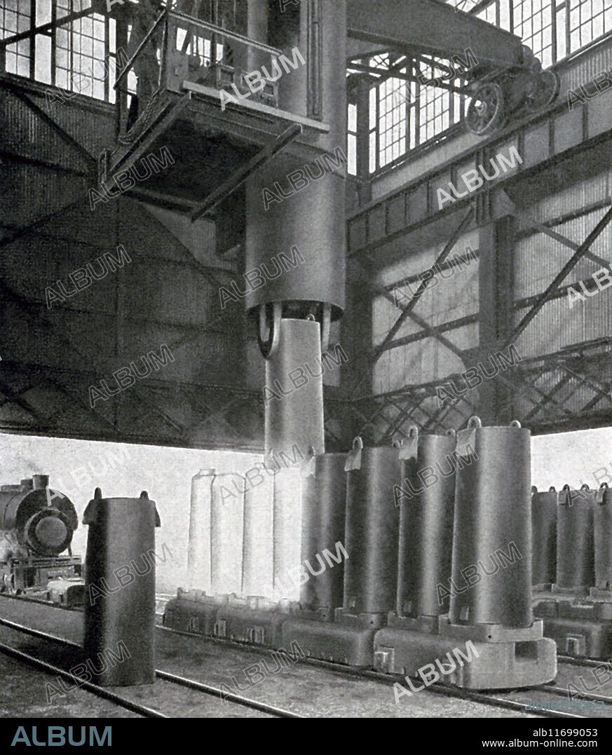 This early 1900s photo shows an Inland Steel Company ingot stripper. The company's business was reducing iron ore to steel. Its only steel mill was located in East Chicago, Indiana, on the Indiana Harbor and Ship Canal and a large landfill protruding out into Lake Michigan. The stripper is a powerful machine that pulls the mold off the ingot. Enormous steel grippers descend and grip the mold near the top, lifting it from the car. Then as steel ram descends and pushes the ingot out through the bottom of the mold, leaving it standing on end, still red hot, on the car passing on to the next mold til the whole row stands stripped and glowing.