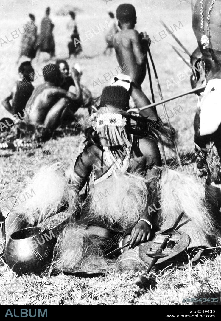 Orig. bildtext... ZULU BRIDAL VEIL THAT IS A MASK. A Zulu bride wearing a leathern and beadwork veil supported by horn hairpins and provided with eyepieces. Guests at the wedding celebrations are taking little notice of her. Anm. Se även BAN171 Field Museum-expeditionen till Angola 1930 Masker Riter Seder och traditioner sites: AFRIKA PhotoDate:1930-01-01.