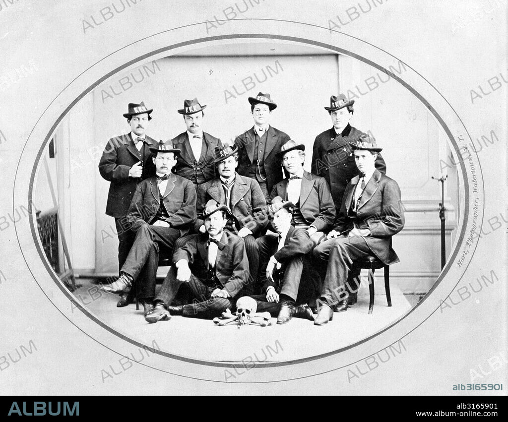 Photograph shows ten men posed, seated and standing, wearing hats with 'KKK' in large letters, and with a skull and bones arranged on the floor in front of them. Ku Klux Klan, Watertown Division, photographer, Watertown, New York.