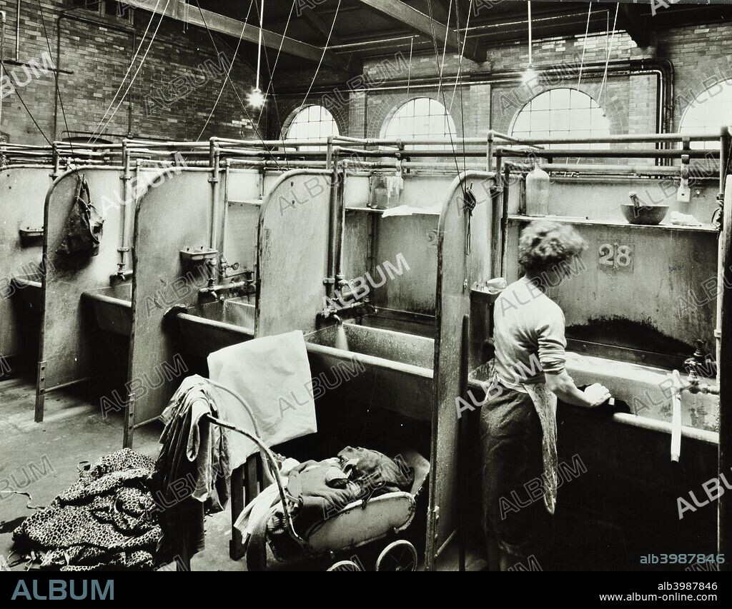 Boundary Street Area Laundry, Bethnal Green, London, 1964. A woman washes some clothes in a deep sink, while more clothes are piled on a pram behind her.