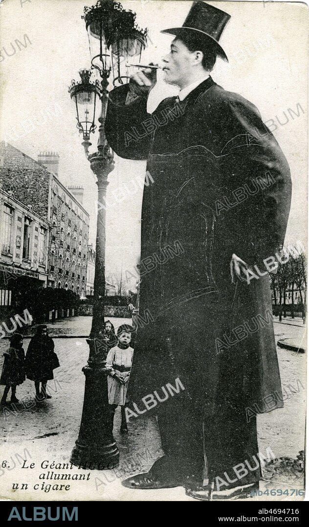 'Le geant Hugo allumant un cigare'. Les freres Hugo presentent un cas de gigantisme unique au monde. Ils sont nes a St Martin Vesubie dans une petite maison isolee sur la montagne. Baptiste l'aine mesure 2.30 et il pese 430 livres. Antoine son frere est ne en 1889, il mesure 2.25 et pese 300 livres, sa taille s'accroit actuellement de 10 centimetres par an. D'apres la faculte de medecine, Antoine Hugo grandira encore plusieurs annees. Les freres Hugo detiennent le record de tous les geants et offrent 1000 francs a leur rival. Carte postale vers 1910, photomontage pour accentuer la taille du geant, par rapport au reverbere. Credit : Jean Vigne/KHARBINE-TAPABOR.