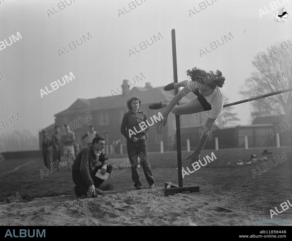 "It's Easy", Says Christine, With An Eye On Olympics. Schoolgirl Christine Wheeler, of Barking Abbey School, Essex who at 16 is a "possible" for Britain's Olympic team, has a 5ft. 6in. mark on the wall at home as an inspiration one day to achieve that height as a high jumper. Now British junior champion at 4ft 11in, Christine is developing her style from the scissors to the western roll - a style which should take her up nearer the mark on the wall and into the Olympic high-jump team. The British women's record is 5ft 3 3/3 ins. Picture shows: Christine Wheeler in action during practice at Motspur Park, London, today (Sunday). Watching her are her coach, Mr A.A. Gold, and Miss Dora Gardener, international high-jumper, who is also a likely candidate for Olympic representation. 14th March 1948.