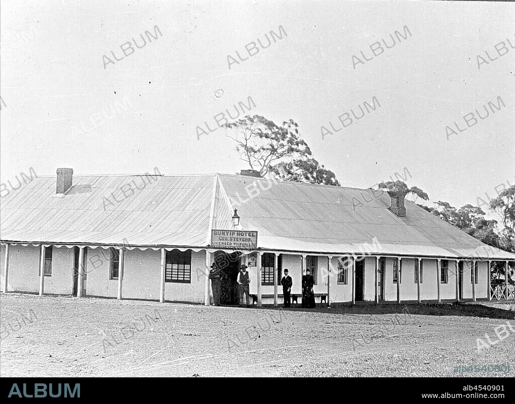 Negative - Group of People Outside Bunyip Hotel, Bunyip, Victoria, circa 1890, A group of people outside the Bunyip Hotel 'Geo. Stevens Licensed Victualler.'. There are two chimneys arising from the building, which appears to have an iron roof and verandah. The people are standing in front of a long seat, and there are many windows across the building. Several large trees can be seen in the distance.