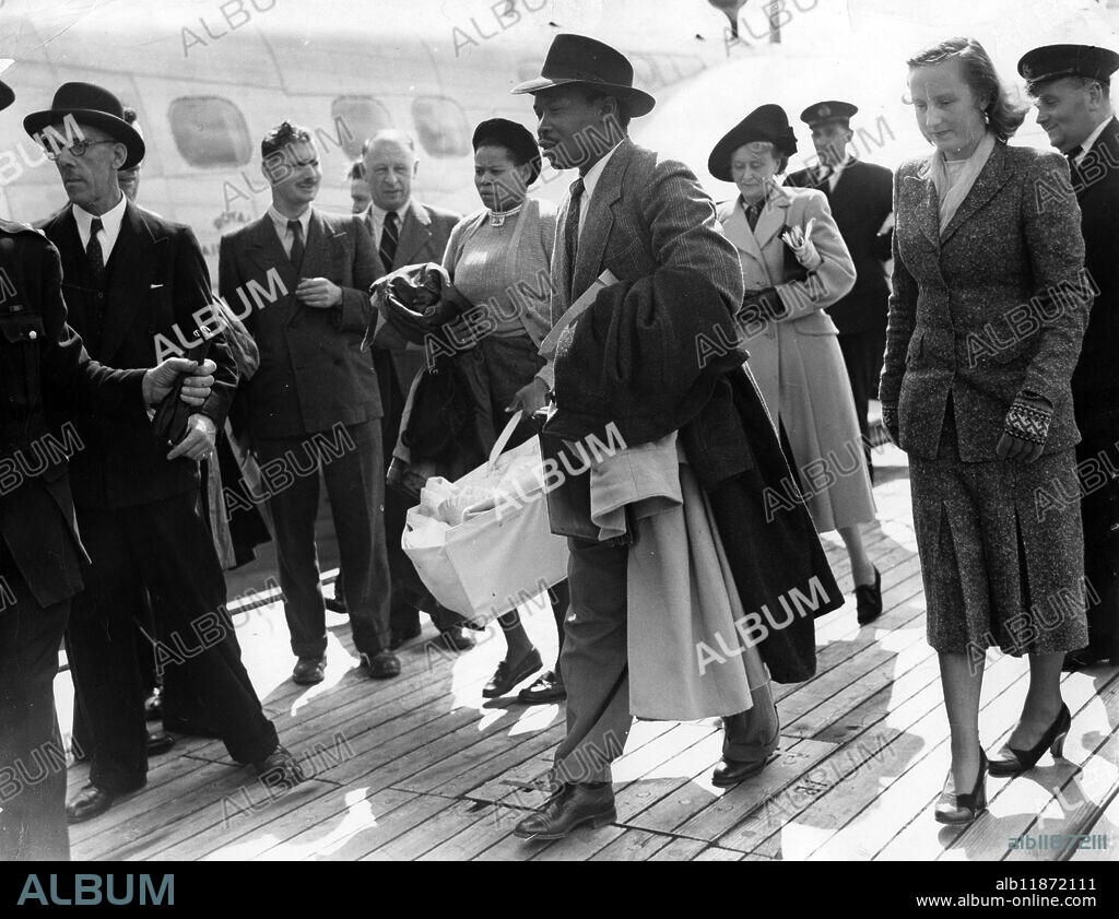 Khamas arrive with daughter. Seretse Khama, exiled Chief designate of the Bamangwato trible of Bechuanaland, his wife a former London typist Ruth Williams, and their three month old daughter Jaqueline arrived at Southampton today by BOAC flying-boat to begin their five years' exile from tribal territory. The Khamas were accompanied by Seretse's sister Nalide. Picture shows: Seretse Khama (centre foreground) carrying Jacqueline in a carry-cot, assisted by Nalide, his sister, on arrival at Southampton today. On right is Mrs Ruth Khama and left of Mrs Khama is seen Mrs Williams (behind) of Lewisham, London, mother of Mrs Khama, who was at Southampton to meet the family. 21 August 1950.