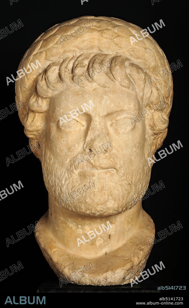 Portrait of a priest or the emperor Diocletian (? ), marble, chiseled, drilled, smoothed, marble, Total: Height: 38 cm; Width: 26 cm; Depth: 26 cm, sculptures, ruler portrait, state portrait, man, head, face, beard, wreath, garland, priest, Diocletian, Diocletian, Roman antiquity, The head, originating from Asia Minor, once belonged to a robe statue, but was worked and inserted separately. It depicts a bearded middle-aged man with staring eyes and a stern facial expression. The voluminous laurel wreath characterizes him as a priest. The physiognomy of the head is determined by broad cheekbones, a powerful jaw and eyes looking into the distance. These are directed upwards and have an incised iris and a pupil that is only flatly depressed. The mouth is drawn down at the sides; with the contracted brows and the nose-mouth folds, a grim mien is created. The hair is shown above the forehead in eight full sickle curls to the left; at the temples and above the ears it is arranged in broad, pointed tufts. Over and behind the ears they are unfinished. The hair mass on the calotte is only given in summary. The thin full beard shows a simple hatching, also the eyebrows. On the head there is a laurel wreath converging above the middle of the forehead and widening from the sides, the leaves of which are regularly offset. Laurel wreaths of the present form are attested for different groups of priests. Such wreaths are also worn by the famous porphyry tetrarchs of San Marco in Venice; a little later the jewel diadem becomes the dominant form. It is no longer possible to determine whether it was a high-ranking military man or official - perhaps a governor - who was portrayed as a member of a college of priests. The design with a thin full beard, strong strands of hair, a strict, unmoving facial expression points to Diocletian's time - occasionally the head is therefore associated with Emperor Diocletian himself.
