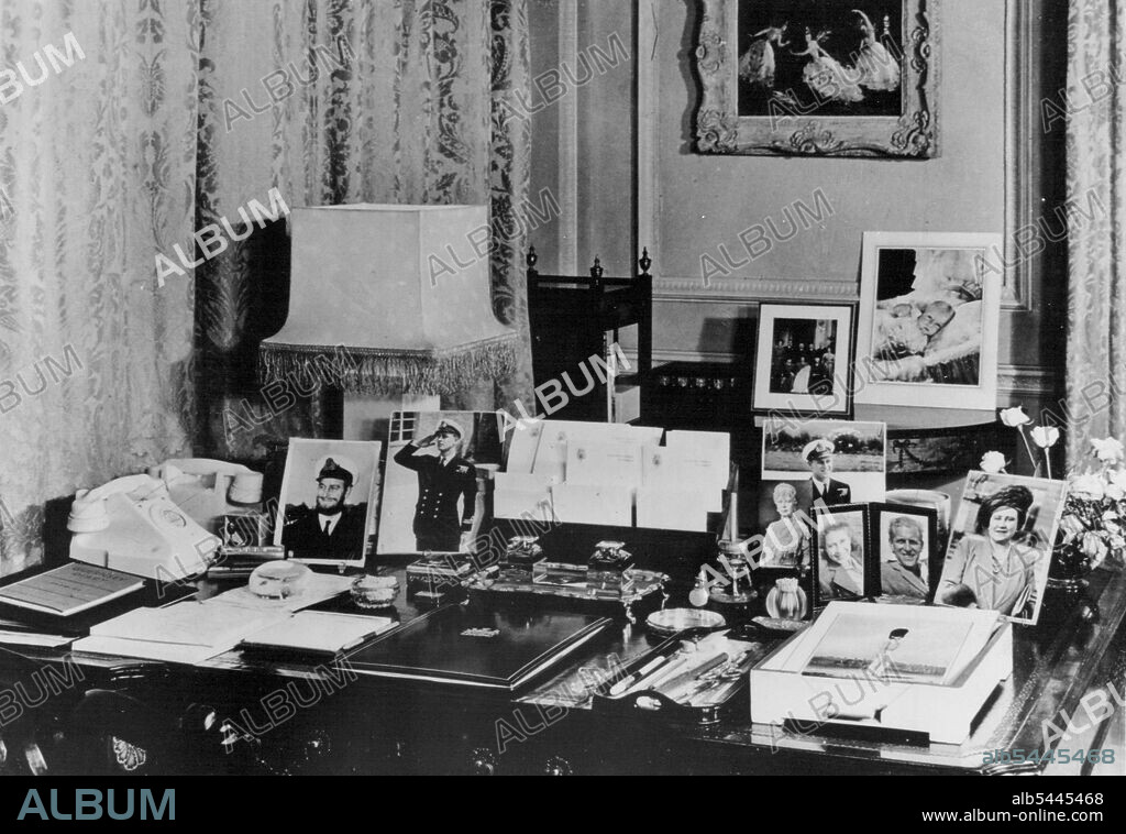 Clarence House - Princess Elizabeth's London Home -- Princess Elizabeth's writing desk in her Sitting Room at Clarence House, the London residence of the Princess and the Duke of Edinburgh. The desk is of the Chippendale period. The photographs are of the Queen, Queen Mary, Princess Margaret and several of the Duke of Edinburgh, including one taken during the war when he wore a beard. Beyond are photographs taken at the christening of Prince Charles, and a painting of the ballet Le Iac des Cygnes, by Oliver Messel. October 28, 1949.