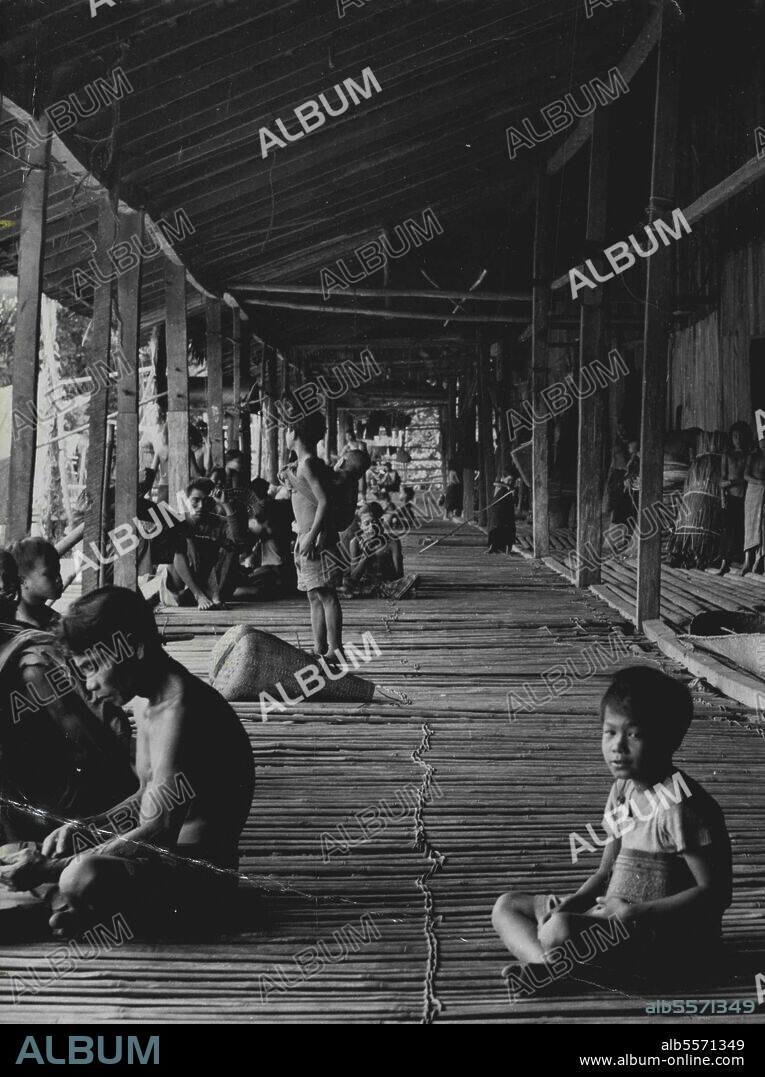Land Dayaks Of Borneo A Dayak Longhouse: The long communal verandah, with the rooms of the individual households leading off from it. Women are pounding rice, and beside the wooden pounders lie winnowing baskets. Groups of men are gossiping or working, a boy is minding his baby brother, and against the wall are bundles of rattan vine for making mats and ropes. Fifty families, comprising up to 400 people, live in houses opening off this long communal veranda where women pound their rice. Rattan for making ropes is piled against the wall. March 8, 1952. (Photo by W.R. Geddes, Camera Press).