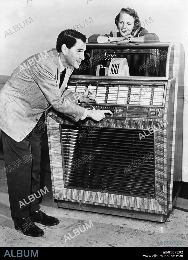 Orig. bildtext... A TOP MUSICAL TRIO Dave Barbour, ace guitarist; Peggy Lee, popular vocalist; and the New Seeburg Select-O-Matic 100. Dave and Peggy, well-known to theater and radio audiences, find their favorite tune on the Select-O-Matic...the new instrument that reproduces their recordings with amazingly life-like tone and quality. This marvelous instrument, just introduced by J.P.Seeburg Corporation of Chicago, gives instant choice of 100 different selections...ranging from popular jazz, old favorites, waltzes and rumbas to the finest of classical music. The Seeburg 100 marks an innovation in the playing of recorded music. Either 10 or 12-inch records may be played vertically without pre-setting. The mechanical brain that is responsible for the amazing performance of the Seeburg works entirely auomatically... with the intriguing and intricate operation in full view. This instrument utilizes the same basic mechanism as the Seeburg Select-O-Matic 200 Library that was widely publicized when introduced early in 1948, and is installed in homes, factories, banks, retail stores, clubs, hotels, restaurants and other commercial organizations throughout the country. Anm. Lyssnare Bildarkivets katalog nr32.6 persons: Peggy Lee ;Dave Barbour sites: USA.