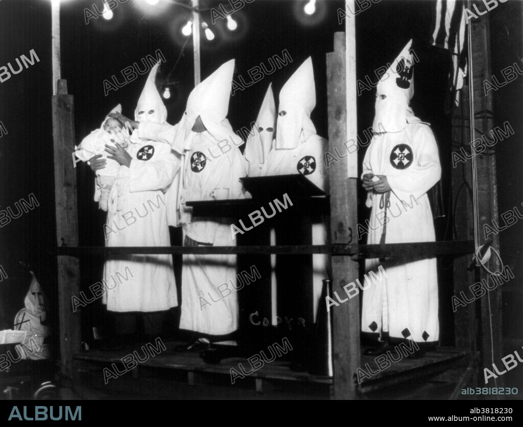 Entitled: "An eight week old child, being Christened by Klansman" shows man holding baby in Ku Klux Klan outfit as another places hand on baby's head; three Klansmen stand in the background on the platform. In 1915 the film The Birth of a Nation was released, mythologizing and glorifying the first Klan and its endeavors. The second KKK was founded in 1915 by William Joseph Simmons at Stone Mountain, outside Atlanta, with fifteen charter members. Its growth was based on a new anti-immigrant, anti-Catholic, prohibitionist and anti-Semitic agenda, which reflected contemporary social tensions, particularly recent immigration. The new organization and chapters adopted regalia featured in The Birth of a Nation; membership was kept secret by wearing masks in public. By the mid-1920s the second Klan had a nationwide reach, with its densest per capita membership in Indiana. The Klan became most prominent in cities with high growth rates between 1910 and 1930, as rural Protestants flocked to jobs in Detroit, and Dayton in the Midwest; and Atlanta, Dallas, Memphis, and Houston in the South. Photographed by Triangle Studio of Photography, 1924.