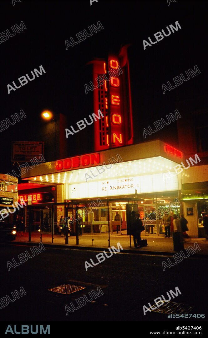 NORMAN WALLEY. Odeon Cinema, New Street, Birmingham, 1985-1987. A view of the Odeon Cinema at night from the north-west, showing the illuminated tower sign and billboard. The final Paramount Theatre, part of a small chain of luxury theatres, opened in 1937 in New Street. The Paramount Theatre was taken over by Odeon in 1942 and its name was subsequently changed. In 1965 the cinema was renovated. In 1988, after this photograph was taken, the cinema was closed for subdivision into six screens. Additional screens were added in 1991 and 1998.