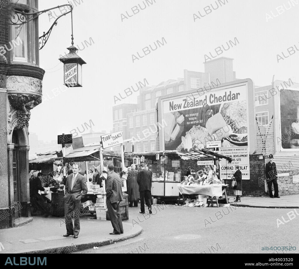 Tubby Isaacs stall, Middlesex Street, Aldgate, London, (1960s?). One of a line of a line of stalls in a street market. A policeman stand on the street corner. Tubby Isaacs' cockels and mussels stall was first established in 1919. The pub on the corner has a lantern outside advertising the brewery Barclay, which was owned by the same family that own Barclay's Bank. They were one of London's most famous breweries, and merged with the brewery Courage in 1955.