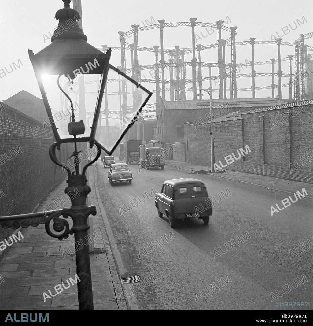 Gas light, King's Cross, London, 1949-1969. A street view with an open gas streetlight in the foreground, two high walls to either side of the road, cars and lorries on the road and the three gasometers at King's Cross in the distance.