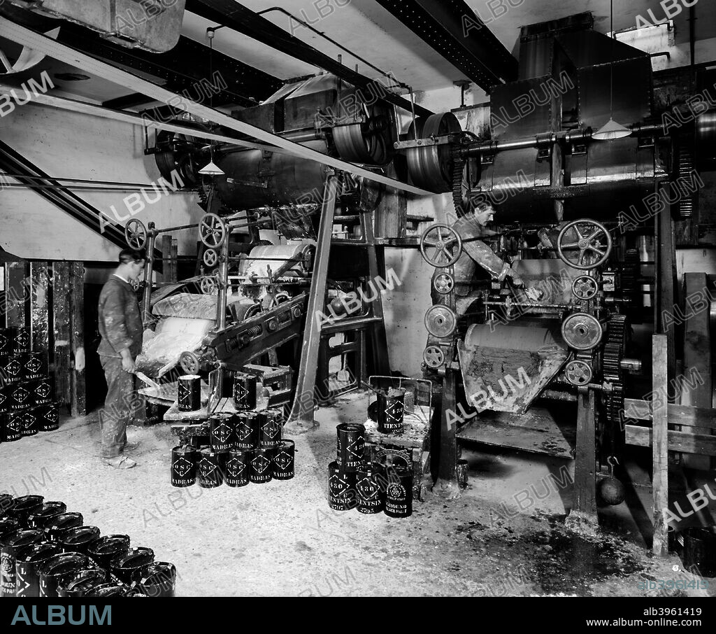 A pair of men at work at Gross Sherwood and Heald's factory, Jenkins Lane, Barking, London, 1910, surrounded by tins of paint marked 'Madras' and 'Tientsin'.