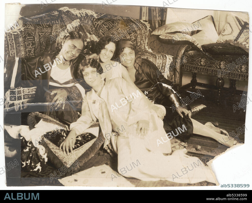 A black-and-white photograph of a group of four (4) young women. The women are sitting on the floor and leaning against a sofa or settee that is draped with a fringed, patterned rug. The woman on the proper left is wearing a kimono-style robe over her garments. The woman in the center of the photograph is wearing a light-colored sash-front dress or blouse and skirt. The third woman is wearing a light-colored sleeveless dress. The fourth woman is wearing a scarf or headwrap on her head, and is wearing a dark colored dress, with her hand lifting her hemline slightly so that her shoes are visible. The two women in the center of the photograph have their hair styled in soft waves. All of the women are smiling and looking at the camera. In the background is another sofa or settee, with several pillows.