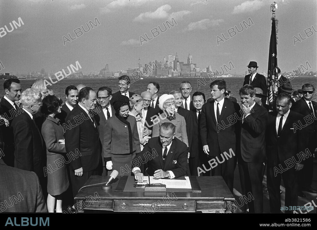 President Johnson signs the Immigration Act as Vice President Hubert Humphrey, Lady Bird Johnson, Muriel Humphrey, Senator Edward (Ted) Kennedy, Senator Robert F. Kennedy, and others look on. Liberty Island, NYC, October 3, 1965. Immigration and Nationality Act of 1965, also known as the Hart-Celler Act, abolished the national origins quota system that was American immigration policy since the 1920s, replacing it with a preference system that focused on immigrants' skills and family relationships with citizens or U.S. residents. The 1965 act marked a radical break from the immigration policies of the past. The law as it stood then excluded Asians and Africans and preferred northern and western Europeans over southern and eastern ones. At the height of the civil rights movement of the 1960s the law was seen as an embarrassment by, among others, President Kennedy, who called the then-quota-system "nearly intolerable". After Kennedy's assassination, President Johnson signed the legislation into law, saying "This system violates the basic principle of American democracy, the principle that values and rewards each man on the basis of his merit as a man. It has been un-American in the highest sense, because it has been untrue to the faith that brought thousands to these shores even before we were a country". Photographed by Yoichi Okamoto October 3, 1965.