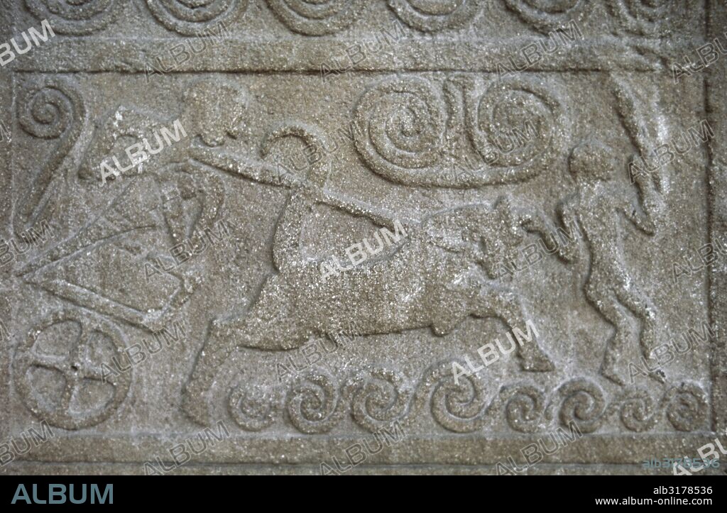 Funerary stele with relief depicting a chariot scene. The standing charioteer pulls at the reins, while a second figure holding a weapon, possibly a sword, is depicted in front of the chariot. The spirals represent waves, indicating the coastal location of the scene. Made in Poros stone. Circle A. Grave V. Mycenae. 1500 BC. National Archaeological Museum. Athens, Greece.