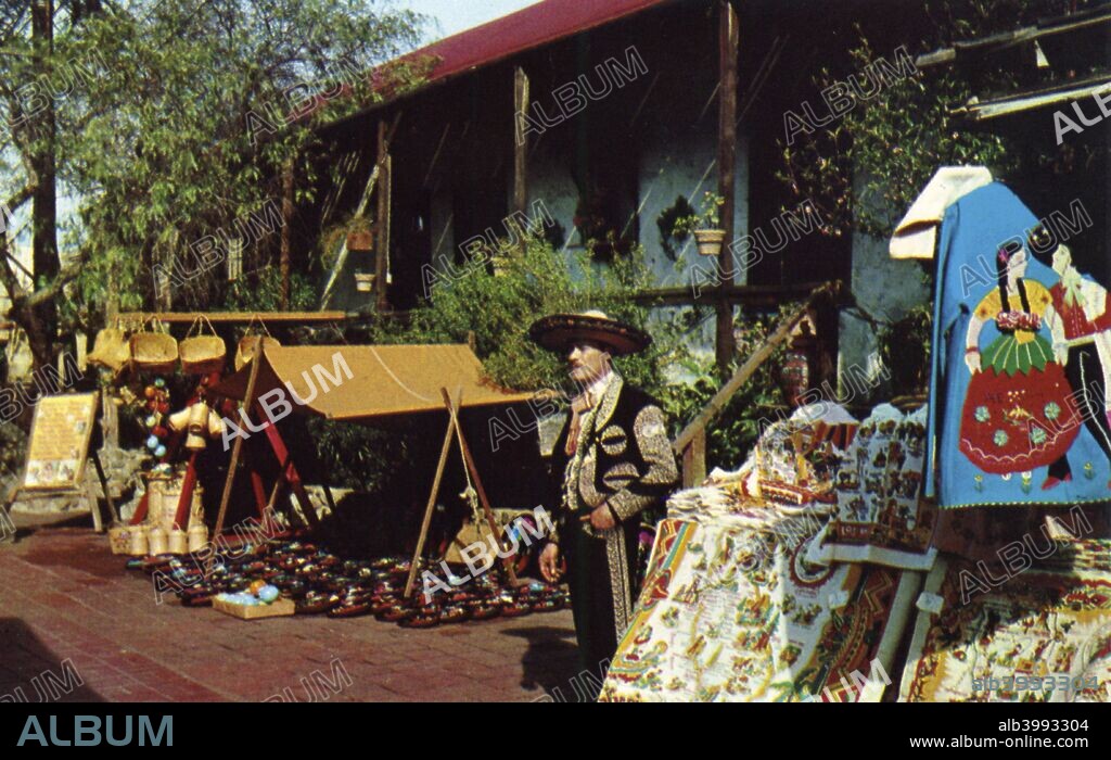 Mexican street merchant, Olvera Street, Los Angeles, California, USA, 1953. Vintage postcard showing a merchant dressed in Mexican garb selling his wares from a lean to in front of the historic Avila Adobe, the oldest standing house in Los Angeles, dating from 1818. Popular with tourists, Olvera Street is a recreation of old Mexico. A Mexican market has been held there since 1930.