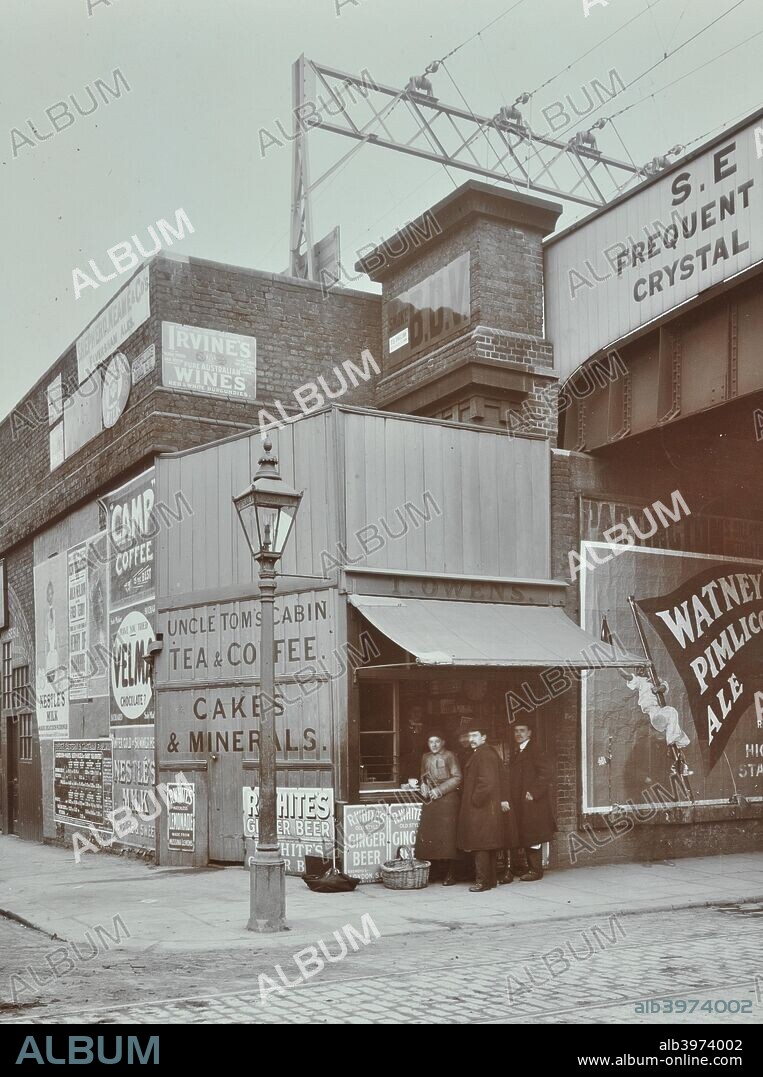 Uncle Tom's Cabin tea stall, 394b Wandsworth Road, London, 1909. Customers stand at a corner tea and coffee stall under the railway bridge. To the left are shops under the railway arches, with a street lamp in front. Signs advertise beer, wine and coffee.
