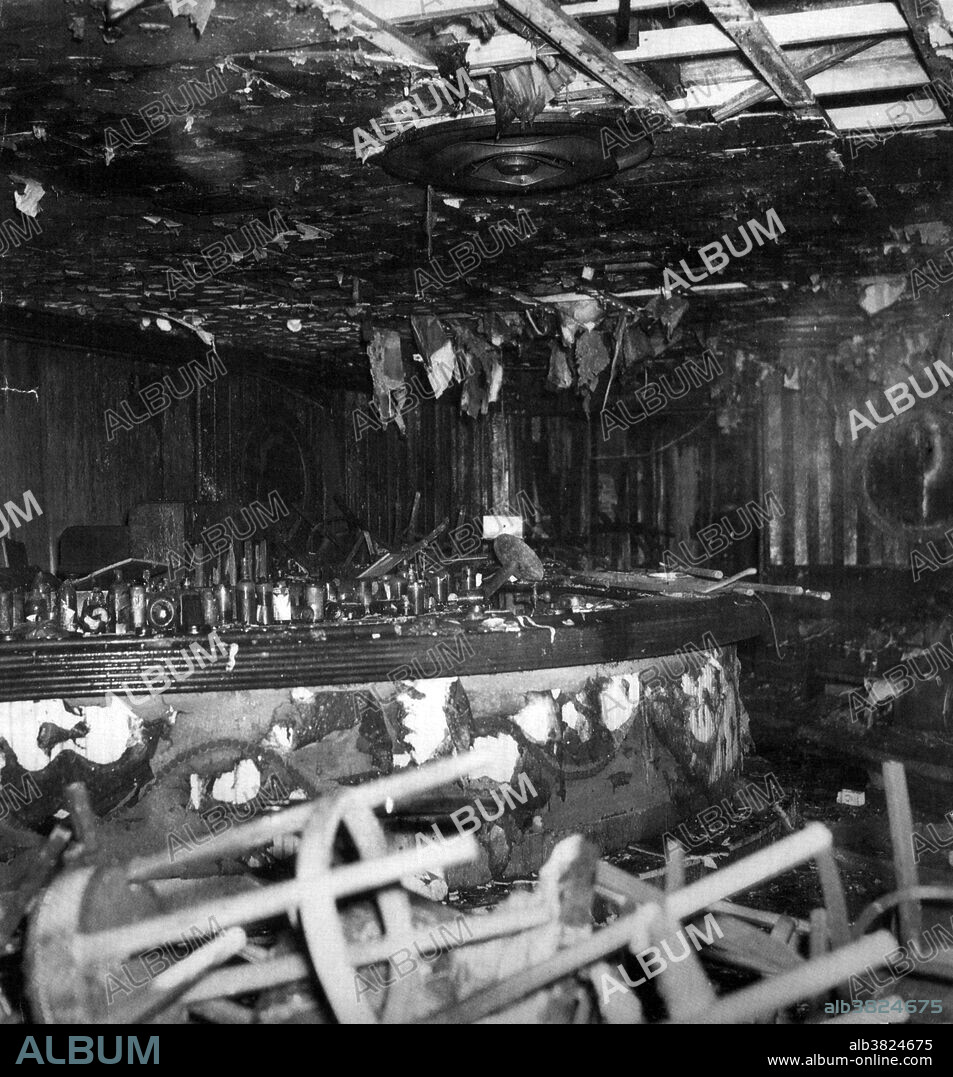On November 28th 1942, a deadly fire occurred at the Cocoanut Grove Nightclub in Boston, Massachusetts, where 492 people perished. The club had a licensed capacity of 500 people, and on the night of the fire there were about 1000 people in the building. At about 10:15PM that evening, a busboy had been ordered to fix a light bulb, but needed to light a match to locate the light socket. Several patrons saw a flicker of a flame in the palm tree of the ceiling decorations. As other furnishings ignited, a fireball of flame and toxic gas raced across the room toward the stairs. The fireball traveled up the stairs and burst into the Foyer area, where coatrooms, restrooms and the main entrance were located. After a small number of people exited, the revolving door became jammed due to the crush of panicked patrons. Observers outside could only watch in horror as relatives and friends were crushed by the weight of the crowd surging against the jammed door. Burnt out interior of the bar of the Cocoanut Grove Nightclub.