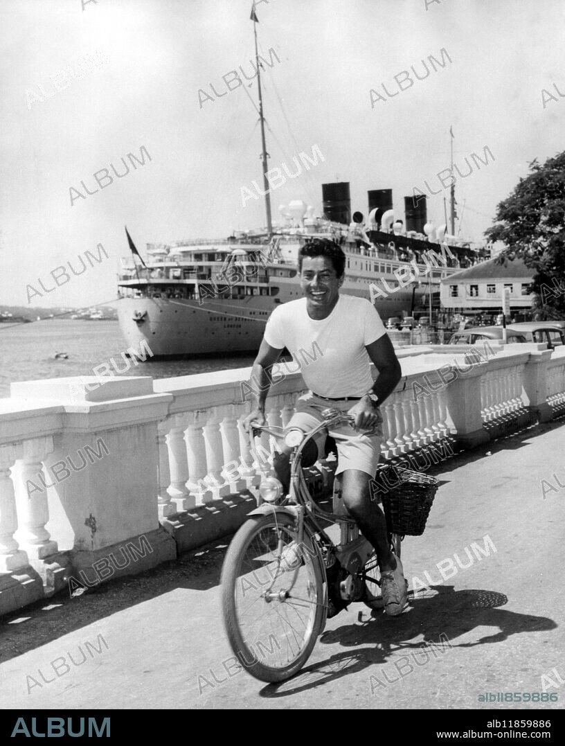 Hollywood actor Farley Granger on Front Street , Hamilton , Bermuda on his motor-assisted bicycle. In the background is the liner Queen of Bermuda . The star was taking a brief holiday in the island sunshine between acting engagements. 13th September 1957.