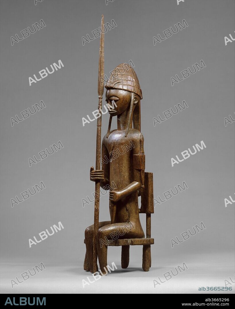 Seated Male Figure with Lance. Culture: Bamana peoples. Dimensions: H. 35 3/8 x W. 9 x D. 10 3/8in. (89.9 x 22.9 x 26.4cm). Date: 16th-19th century.
Bamana figures such as this were the focal point of celebrations of the "Jo" and "Gwan" initiation societies (associations of men and women) in several villages in southern Mali. This work depicts an idealized male leader that would have been the companion to a similarly attired mother and child representation. For annual rituals, a community's collection of a series of such works were removed from shrines, cleaned and oiled, decorated with cloth and beads, and displayed in the village square as a group. The mother and child and her male counterpart were placed in central positions of honor, distinguished by attributes of their extraordinary physical and supernatural powers.
This figure is depicted wearing a hunter's hat adorned with animal horns and amulets. The amulets serve to heighten his powers of perception and in doing so augment his abilities to find game. He holds a lance considered a "weapon of distinction." Such arms, relics of heroic ancestors, were handed down from father to son at the time of initiation. The knife worn on his upper arm is also associated with hunting and is of the type frequently seen on older examples of Bamana sculpture, including terracottas dating as far back as the twelfth century. The heavily lidded eyes, closed mouth, and arms held close to the body suggest a sense of calm and self-possession, accentuating the stature and respect commanded by the figure.